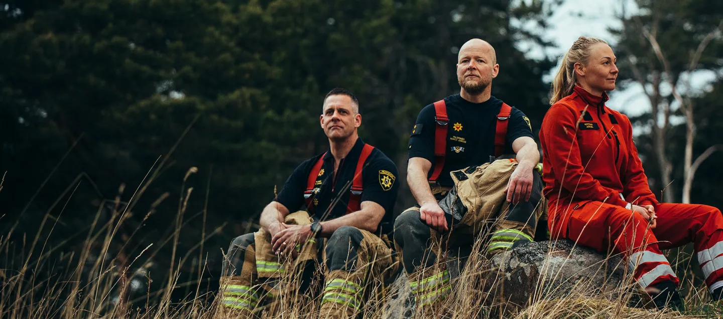 Kolme pelastustyöntekijää istuu pellolla metsän laidalla.