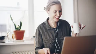 Nainen kannettavan tietokoneen äärellä nauramassa ja kahvikuppi kädessä