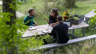 Kolme henkilöä istuu piknikpöydän ympärillä vehreässä ympäristössä. 